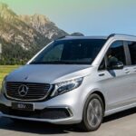 A silver Mercedes-Benz EQV 7-seater electric van driving on a scenic road with a background of green fields and rocky mountains under a blue sky.