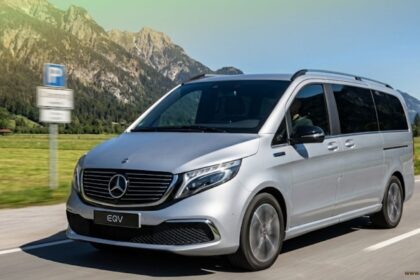 A silver Mercedes-Benz EQV 7-seater electric van driving on a scenic road with a background of green fields and rocky mountains under a blue sky.