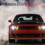 A red Dodge Challenger SRT Demon performing a burnout on a drag strip at night, with thick white smoke billowing from behind the car and a racing tree light visible in the background.
