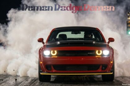 A red Dodge Challenger SRT Demon performing a burnout on a drag strip at night, with thick white smoke billowing from behind the car and a racing tree light visible in the background.
