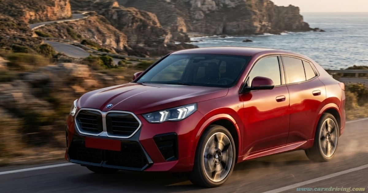 Front three-quarter view of a metallic red BMW iX2 electric coupe SUV driving along a scenic coastal road at sunset.