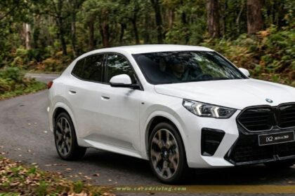 A white BMW iX2 xDrive30 electric coupe SUV driving on a winding, asphalt road surrounded by a dense green forest. The car is shown from a three-quarter front angle, highlighting its black kidney grille, sleek headlights, and sporty design.