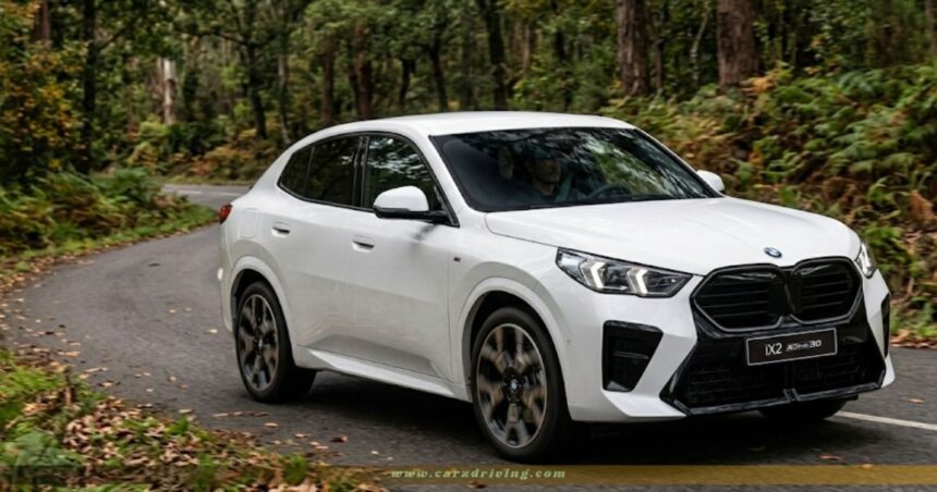 A white BMW iX2 xDrive30 electric coupe SUV driving on a winding, asphalt road surrounded by a dense green forest. The car is shown from a three-quarter front angle, highlighting its black kidney grille, sleek headlights, and sporty design.