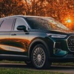 A dark green Chery Tiggo 9 SUV parked on a roadside path during a vibrant golden hour sunset, with trees and a brick wall in the background.