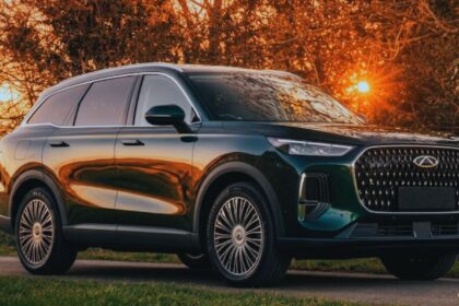 A dark green Chery Tiggo 9 SUV parked on a roadside path during a vibrant golden hour sunset, with trees and a brick wall in the background.