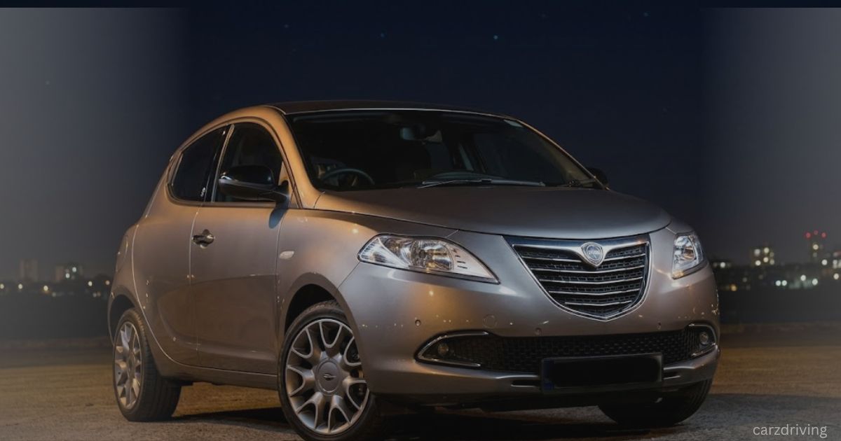 Front three-quarter view of a silver Chrysler Ypsilon hatchback parked outdoors at night under a dark sky with city lights in the blurred background.