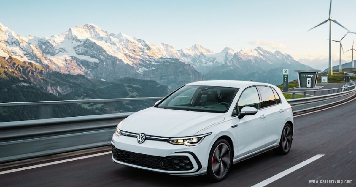 A white Volkswagen Golf GTE driving on a scenic mountain road with snow-capped peaks in the background.