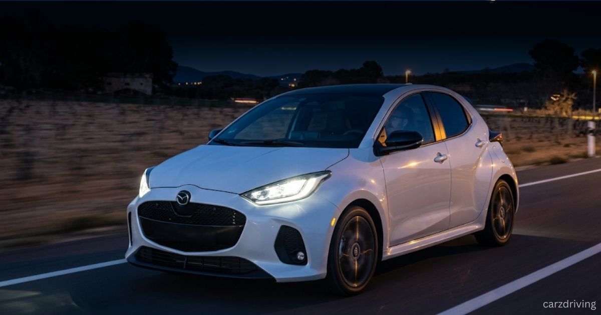 A white Mazda 2 Hybrid hatchback driving on a road at dusk with its headlights illuminated.