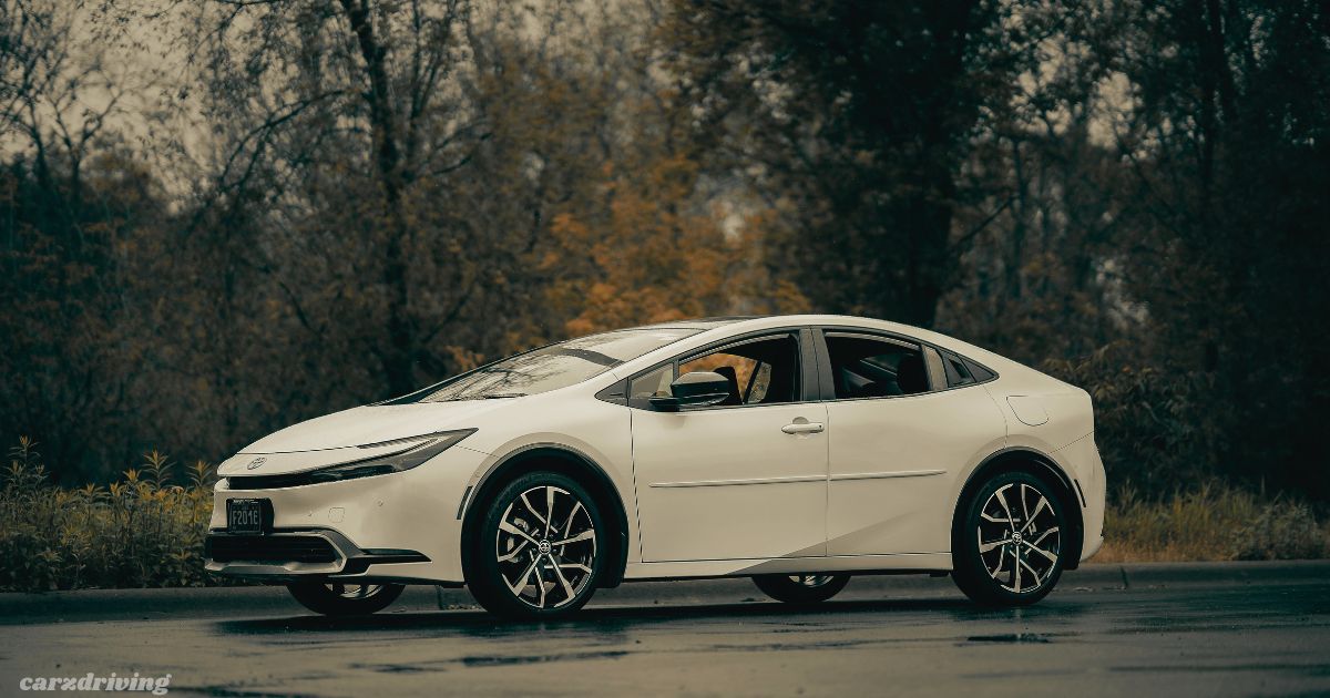 A white fifth-generation Toyota Prius sedan parked on a damp road, shown from a three-quarter side angle. The car features distinct multi-spoke alloy wheels and sleek headlights. The background is filled with blurred, dark trees showing muted autumn colors, and a "carzdriving" watermark is visible in the bottom left corner.