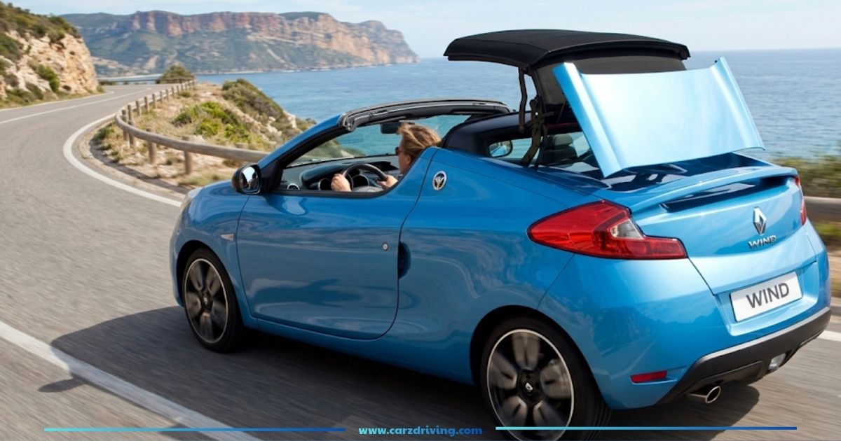 A metallic blue Renault Wind convertible driving along a scenic coastal road with its unique hardtop roof mid-way through pivoting open.