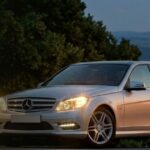 Silver Mercedes-Benz W204 sedan with headlights on at dusk.