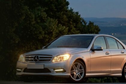 Silver Mercedes-Benz W204 sedan with headlights on at dusk.
