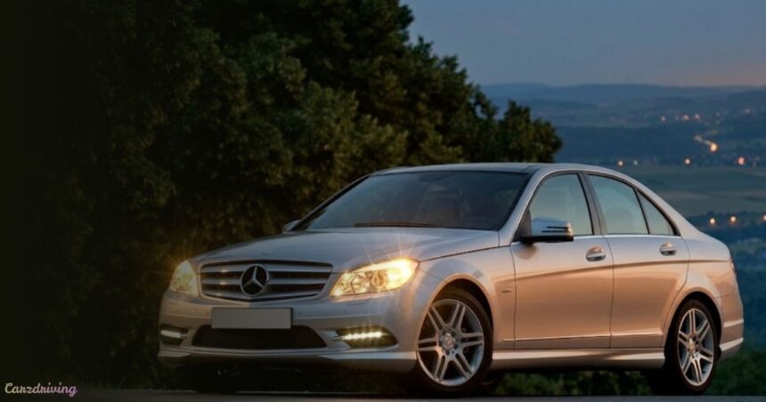 Silver Mercedes-Benz W204 sedan with headlights on at dusk.
