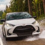 "White Toyota Yaris GR Sport hatchback splashing through a puddle on a wet forest road"