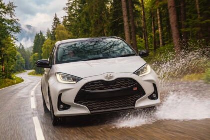 "White Toyota Yaris GR Sport hatchback splashing through a puddle on a wet forest road"
