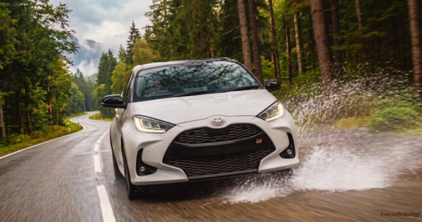 "White Toyota Yaris GR Sport hatchback splashing through a puddle on a wet forest road"