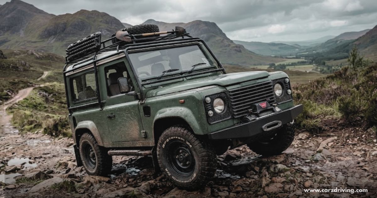 Classic green Land Rover Def 90 off-roading on a rocky mountain trail with a roof rack and recovery gear