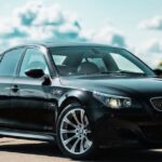 A black BMW E60 5 Series sedan parked on a roadside with a blurred natural background of water and trees.