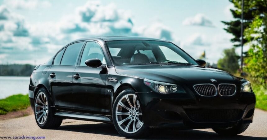 A black BMW E60 5 Series sedan parked on a roadside with a blurred natural background of water and trees.