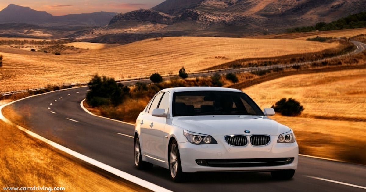 A white BMW E60 5 Series sedan driving along a winding road through a scenic, golden-hued hilly landscape.