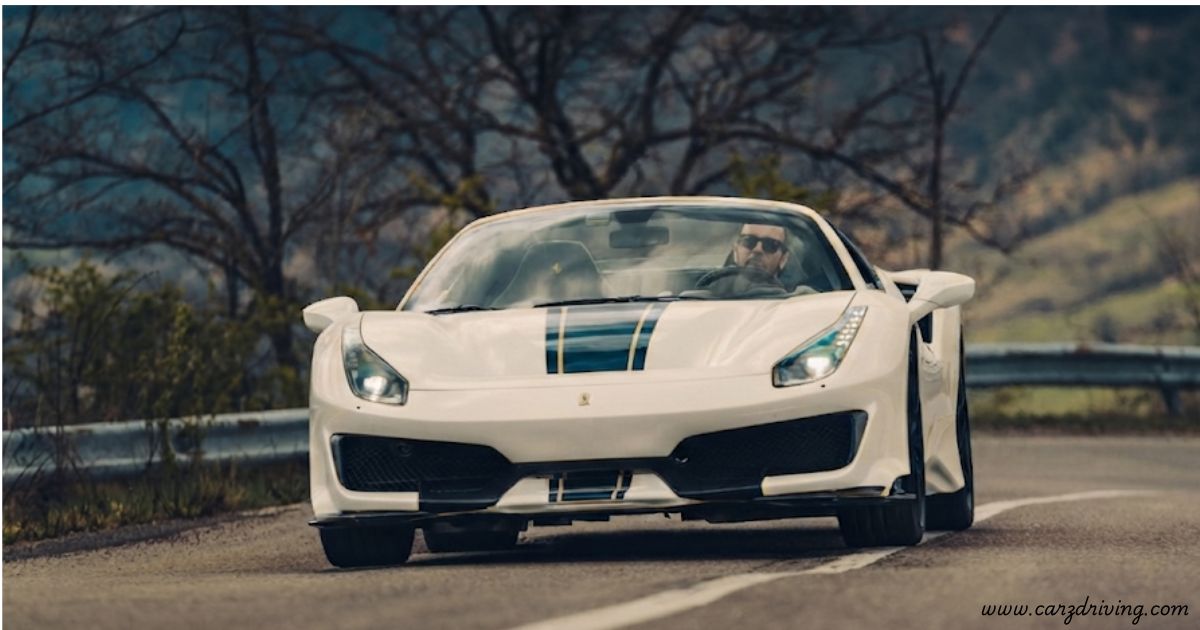 "Front view of a white Ferrari 488 Pista driving on a winding countryside road, featuring a prominent blue and gold racing stripe, with bare trees in the background."