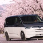 "A front-three-quarter view of a two-tone white and grey VW Multivan driving along a winding road lined with blossoming cherry blossom trees."