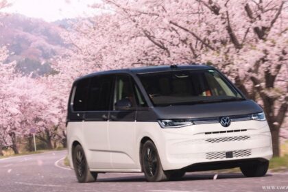 "A front-three-quarter view of a two-tone white and grey VW Multivan driving along a winding road lined with blossoming cherry blossom trees."