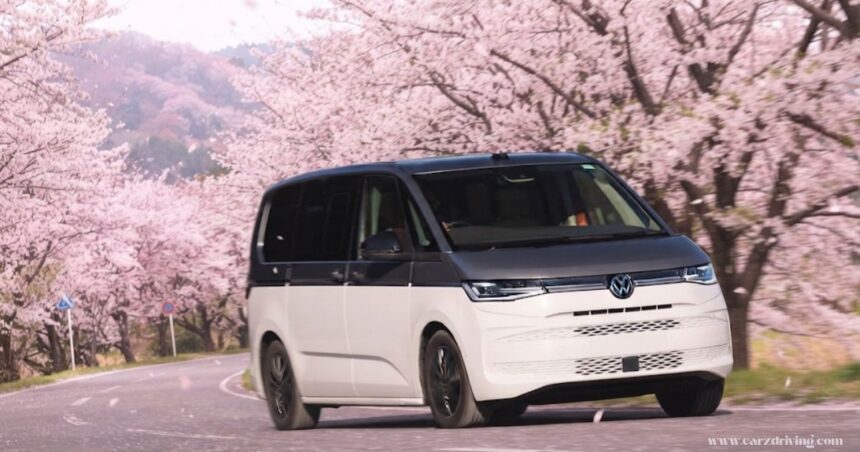 "A front-three-quarter view of a two-tone white and grey VW Multivan driving along a winding road lined with blossoming cherry blossom trees."