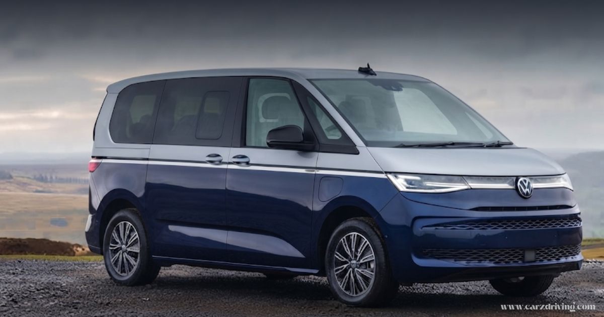 "A side-profile view of a two-tone blue and silver VW Multivan parked on a gravel surface against a cloudy, mountainous backdrop."