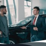 A car salesman holding a clipboard and pen talking to a young couple inside a car dealership next to a dark sedan, with text in the upper left reading "Car Finance Eligibility Checker."
