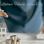 An image with the text "Labour House Value Tax" featuring a small model house surrounded by stacks of coins and paper money on a desk, with a person's hands hovering above the house and using a calculator in the background.