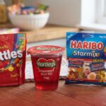 An assortment of packaged sweets with least calories displayed on a wooden surface, including a red bag of Skittles Fruits, a Hartley's strawberry jelly pot, a blue bag of Haribo Starmix Minis, and three Chupa Chups lollipops in a glass jar.