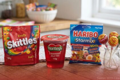 An assortment of packaged sweets with least calories displayed on a wooden surface, including a red bag of Skittles Fruits, a Hartley's strawberry jelly pot, a blue bag of Haribo Starmix Minis, and three Chupa Chups lollipops in a glass jar.