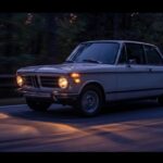 Vintage BMW 2002 cars sedan with headlights turned on driving along a quiet road at twilight.