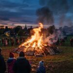 A large bonfires before 7pm uk burning outdoors at dusk surrounded by a crowd of people.
