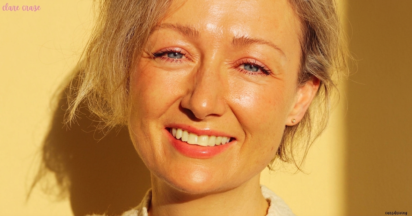 Close-up portrait of Clare Craze smiling softly against a warm yellow background illuminated by sunlight.