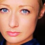 A close-up headshot of clare craze fair-skinned woman with short blonde hair and striking blue eyes, facing forward with a neutral expression and light pink lipstick.