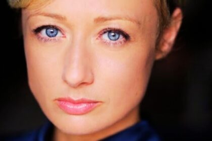 A close-up headshot of clare craze fair-skinned woman with short blonde hair and striking blue eyes, facing forward with a neutral expression and light pink lipstick.