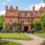 **Alt Text:** Students in academic gowns walk along a stone pathway outside the red-brick building of Girton College, Cambridge, surrounded by manicured lawns, green trees, and purple wisteria representinf one of the list of cambridge colleges