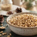 A ceramic bowl filled with raw pine nut nuts on a table next to pinecones.