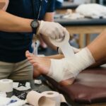 Close-up of a healthcare provider applying athletic taping and bandaging to a patient's foot and lower leg in a clinical setting, with supplies on the table.