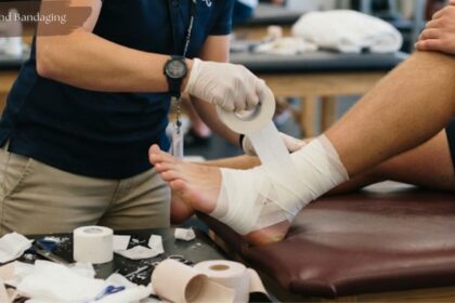 Close-up of a healthcare provider applying athletic taping and bandaging to a patient's foot and lower leg in a clinical setting, with supplies on the table.