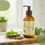 Amber glass pump bottle of Botanical Essence Tea Oil Face Wash surrounded by tea leaves and botanical seeds on a wooden counter.