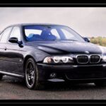 Front-three-quarter view of a dark metallic BMW E39 M5 sedan with iconic halo headlights parked outdoors.