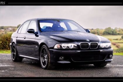 Front-three-quarter view of a dark metallic BMW E39 M5 sedan with iconic halo headlights parked outdoors.