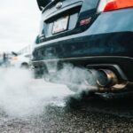 Rear view of a dark grey Subaru STI parked on wet pavement with exhaust smoke billowing from its dual exhaust pipes.