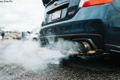 Rear view of a dark grey Subaru STI parked on wet pavement with exhaust smoke billowing from its dual exhaust pipes.