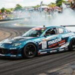 Mazda RX-8 drift car sliding around a track corner, generating thick white tire smoke and flames from the exhaust while spectators watch from the background.