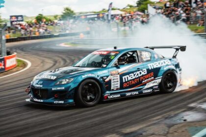 Mazda RX-8 drift car sliding around a track corner, generating thick white tire smoke and flames from the exhaust while spectators watch from the background.