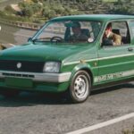 A vintage green MG Metro hatchback driving along a winding coastal road, featuring white side striping with "MG METRO" text, a rear spoiler, and overlay text reading "MG METRO" in the top-left corner.